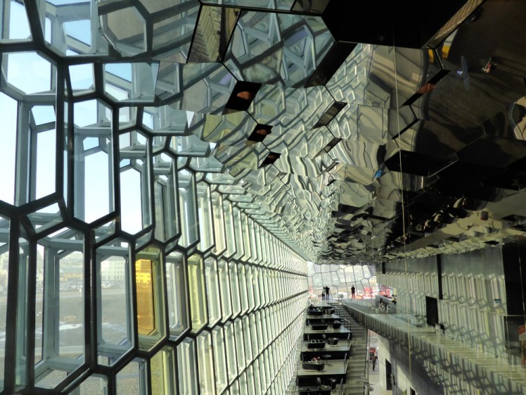 A photo from upstairs inside Harpa. The whole thing is a slightly dizzying array of hexagonal glass panes with some open stairs in the middle. The glass is slightly green-tinted with a few panes in shades of yellow.