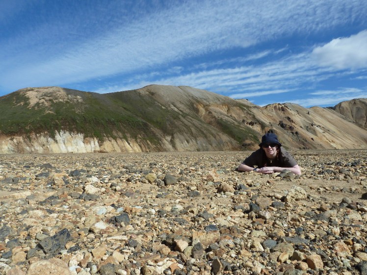 Me lying in a gravel field looking at the camera. Behind me are yellow striped mountains with thick dark green moss growing on them.