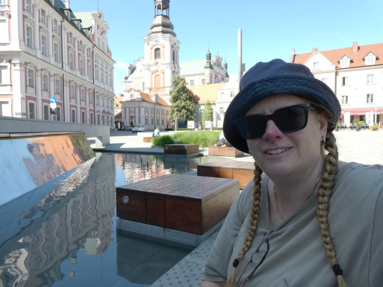 Selfie in Plac Kolegiacki, with pink and white buildings behind me, a water feature in front of me. I'm sitting in the shade but behind me is sunshine. I'm wearing my grey-green Taylor Swift reputation tour t-shirt and my usual plaits.