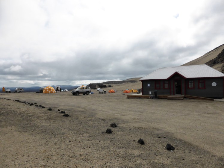 Dreki, a large green mountain hut. Beside it are lots of yellow and orange tents. The landscape is greyish yellow - pumice erupted out of the volcano that you can't quite see to the right of the picture.