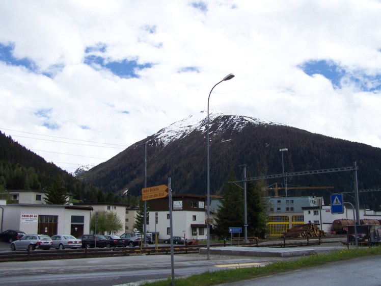 Davos in summer. A snow-capped mountain looms over a very ordinary and industrial-looking town and main road.