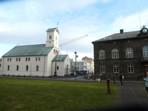 Iceland's Parliament Square in Reykjavik. To the left is a fairly small white church with a green roof - this is Reykjavik Cathedral. To the right is a two-storey building of dark stone with lots of arch-shaped windows. This is Parliament House. Behind the two is a very prominent crane that might be building the extension.