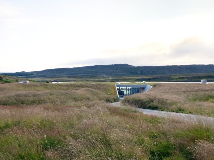 A grassy field with a turf-roofed building set into the low hill. The only sign it's there is the concrete walls supporting the sides and the glass doors. Above the hill, you can see a sliver of lake between the field and the mountain beyond.