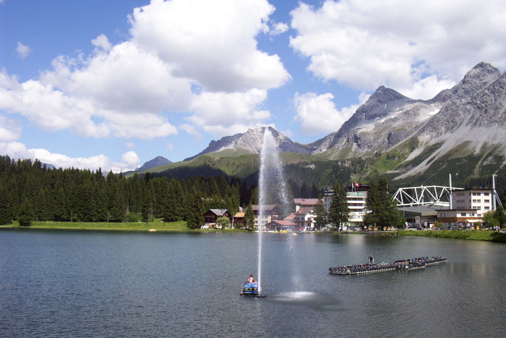 A fountain jetting up in the middle of a small lake. On the right-hand shore are a cluster of wooden chalets and more modern buildings. On the left-hand shore is pine woodland. Behind everything are craggy mountains. The village is just below the treeline. The sky is blue and cloudy.