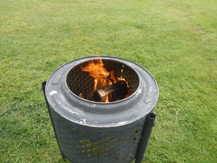 A fire burning merrily in a steel barrel-shaped fire pit in a green field.