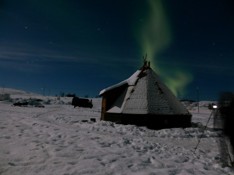 A Sami lavu, a pyramid-shaped tent, in the snow with the Northern Lights apparently bursting out from behind it.