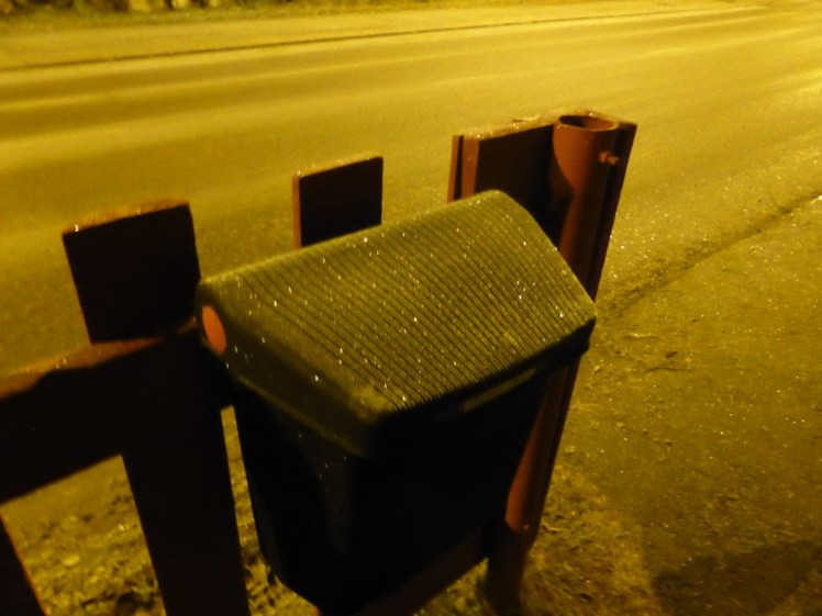 A bin with a lid mounted on a wooden fence. Everything is lightly sprinkled with snow and sparkles under the yellow light of a streetlamp. This represents the time I got on the wrong train and arrived very late in the Arctic.