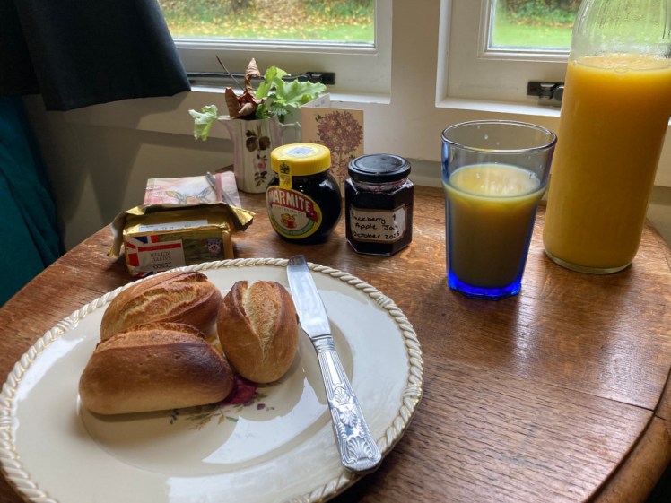 Breakfast laid out on the tiny table - a vintage plate and beaded knife with some baked rolls, a block of butter, jar of Marmite & jam, a blue soda glass of orange juice and a glass bottle with the rest of the juice, all on a fold-out table with a hint of garden out the window behind it.