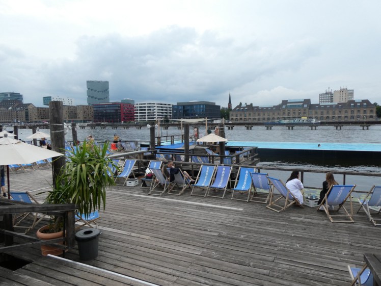 Two levels of decking leading down to a long thin pool floating in the river. There are deckchairs on each of the decks and industrial Berlin visible on the other side of the river.