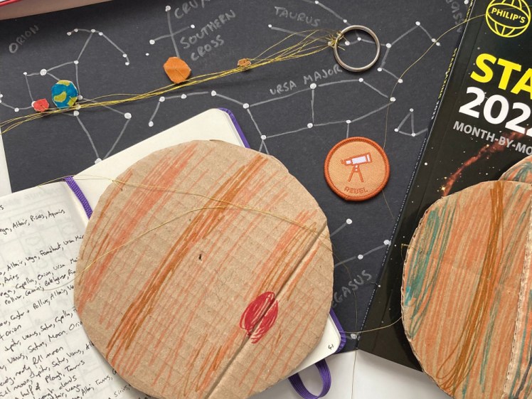 My Stargazing stuff - a notebook with my star log scribbled in, my model solar system, my stargazing book and my Rebel Stargazing badge, which is a small brown sew-on badge with a telescope on it.