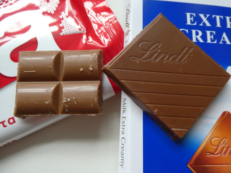A piece of each of Lindt & Lacta chocolate sitting on top of their packaging next to each other so you can see that the Lacta is a much lighter colour than Lindt.