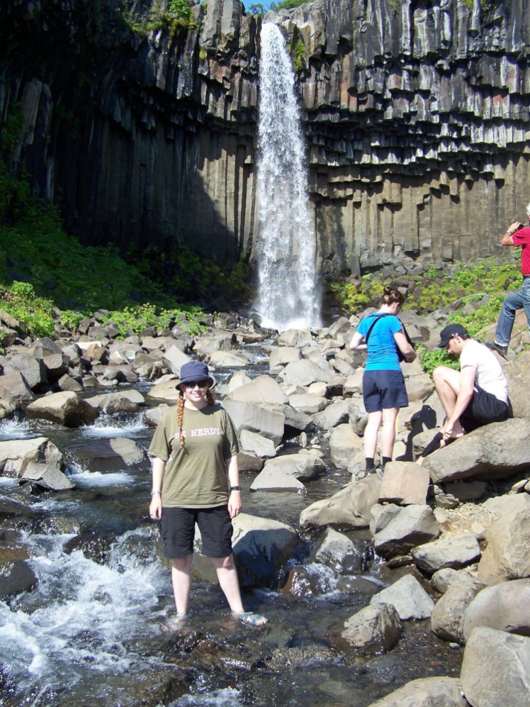 Another timer selfie. I'm in Iceland, standing in the stream below a narrow but pretty waterfall that's falling through a low cliff of basalt columns. I'm wearing mountain sandals in light blue and you can hardly see them in the water, black shorts and an olive green t-shirt.