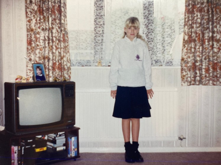 Me, aged 11, in my new secondary school uniform in a house that looks decidedly 80s, although it's September 1996. I'm wearing a sweatshirt so pale grey it almost looks white over a white shirt, a black knee-length skirt, dark socks with chunky 90s school shoes.