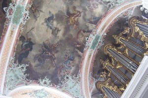 The ceiling of St Gallen Cathedral. Most of the dome is filled with a fresco in muted colours, surrounded by arches in pink and gold, with green corners and plaques. You can also see the organ pipes, which are decorated in gold.