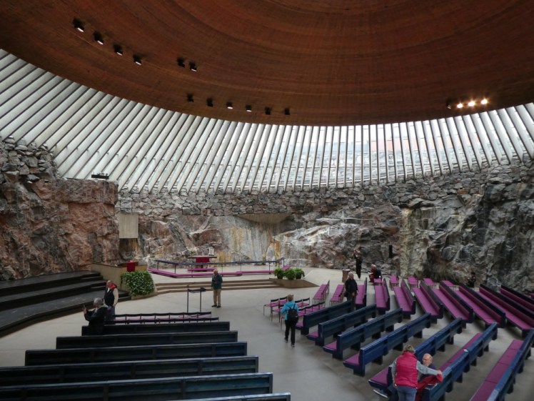 Inside the Church in the Rock. The copper dome is golden-brown inside, held up by concrete struts which let light in between. The walls below the concrete are rough rock and then the floor of the church itself looks like your average modern church.