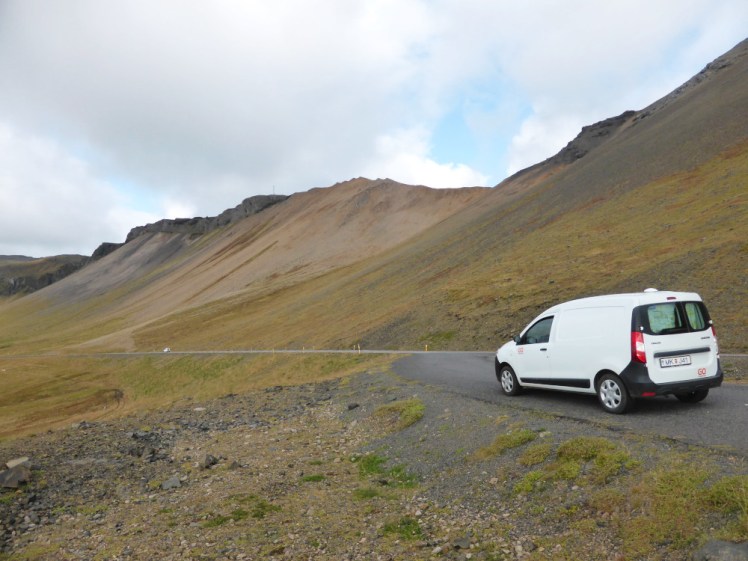 A small panel van campervan parked on the side of a road in Iceland which has been cut into the side of a shallow-sloped mountain, covered in yellow-green moss.
