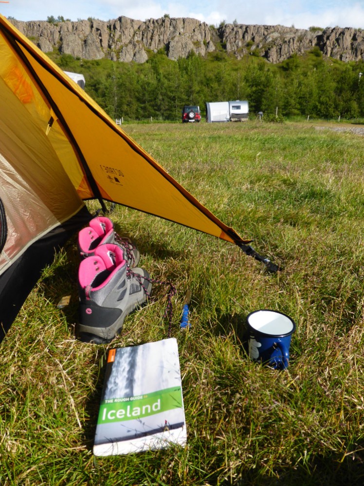 The tiny porch of my tent with a pair of boots, an enamel mug and a guidebook in it. Behind the tent is a row of long low inland lava cliffs.