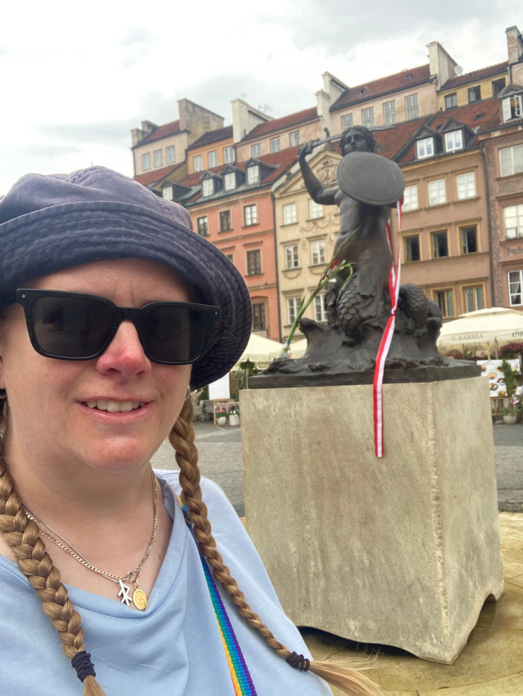A selfie with the Warsaw Mermaid statue in the middle of a fountain. I'm wearing a light blue t-shirt and a navy bucket hat with the brim folded back. The Mermaid is holding a round shield and a sword and has a length of red and white ribbon hanging from her.