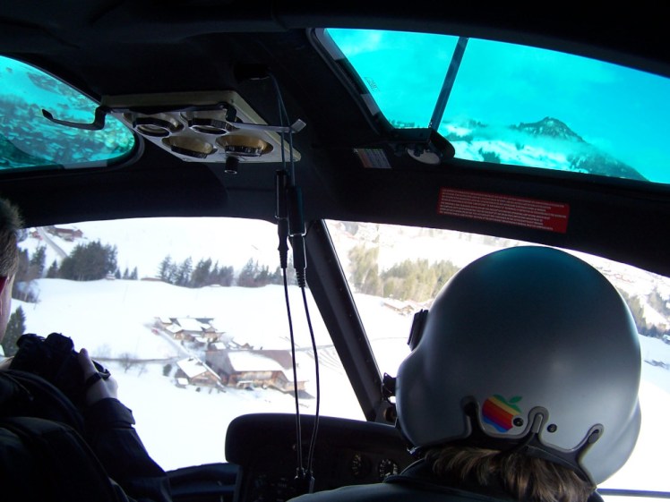 The back of our pilot's head, wearing a white helmet with a rainbow Apple sticker on the back. You can see out the front windows of the helicopter to a village in the snow.