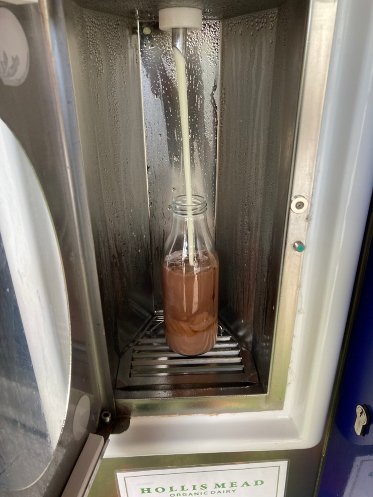 A glass milk bottle with an inch of chocolate syrup in the bottom being filled with milk from a vending machine dispenser.