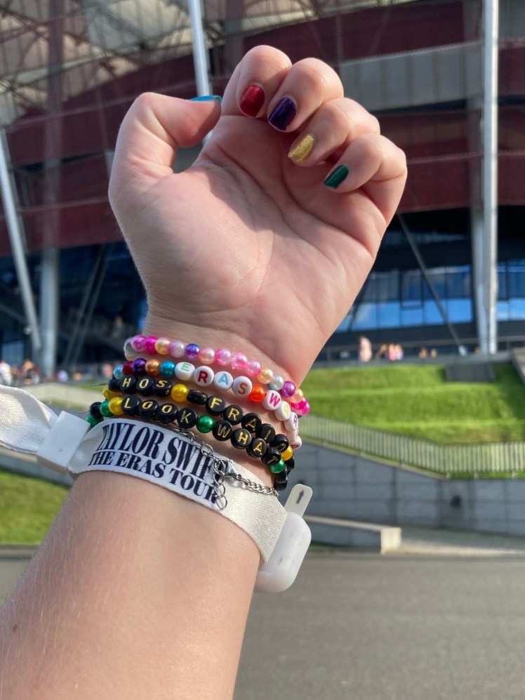 My hand held up in front of the stadium to show off my light-up tour wristband, my friendship bracelets and my Eras nails, Taylor Swift to 1989 (Reputation to Midnights is on the other hand).