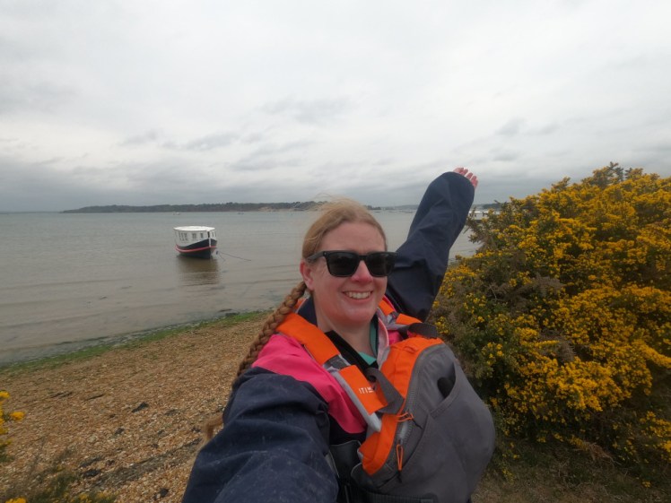 Standing with my arm raised in front of a patch of grey sea, wetsuit hidden by a navy & pink sailing cagoule and an orange & grey buoyancy aid.