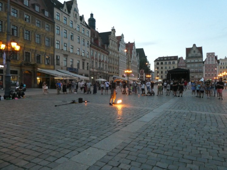 A fire performer, holding up a ball of flame on a rope, surrounded by crowds in the half-darkness of the square.