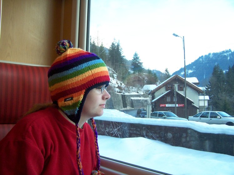 Me, sitting on a train wearing a round-necked red fleece jumper and a rainbow-striped hat with earflaps, looking out of the window at a village under a nice blanket of snow.