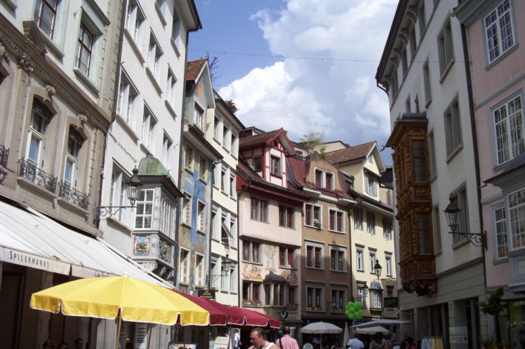 A narrow street in St Gallen, with tall four or five storey houses and pointed roofs. At ground level, there are umbrellas outside cafes and restaurants.