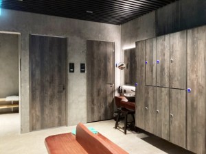 The changing rooms. The lockers look like grey weathered wood or very raw concrete. The floor and walls are white stone and there's a bench in the middle in an oddly bright orange shade of wood.