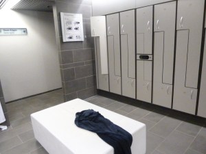 The Blue Lagoon changing rooms. Each vertical space is divided into two lockers with a squarish area for a bag on either top or bottom and a long bit for hanging coats. In the middle is a white seat with a wet towel on it. The whole room is in shaedes of grey with polished stone walls.