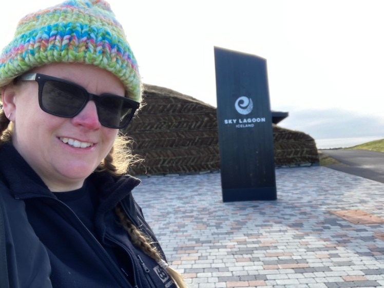 Selfie outside the Sky Lagoon. The turf wall and the big SKY LAGOON sign are both visible behind me. I'm wearing a black jacket, sunglasses and a knitted had that manages to be pastel and neon at the same time.