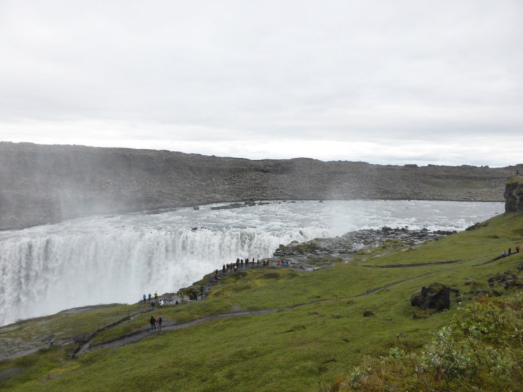 Dettifoss, a huge waterfall falling into a canyon and out of frame. It still manages to dwarf both banks and the line of tourists taking photos of it.