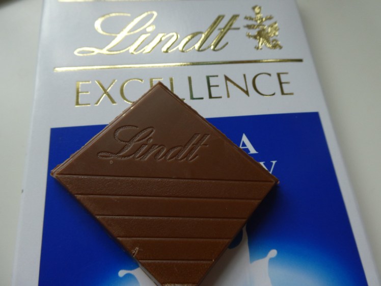 A square of Lindt Excellence chocolate lying on top of its blue and white box, with the letters of its name shining in gold foil.