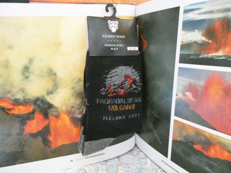 A pair of black socks with grey heels and toes. An image of a volcano erupting against a full moon is embroidered on them, along with the words "FAGRADALSFJALL VOLCANO ICELAND 2021"