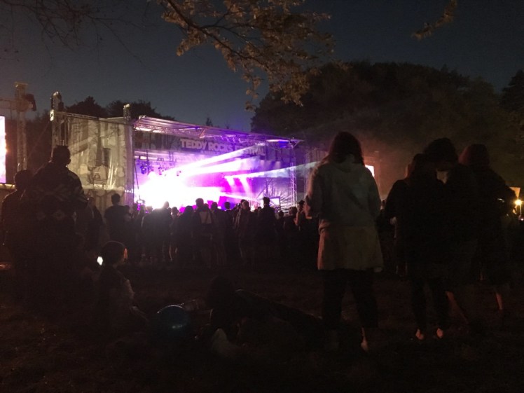 The main stage at Teddy Rocks 2018, seen from near the back of the small field where it stands. Purple lights are shining from it and I guess Ash must be on the stage but you can't see them.