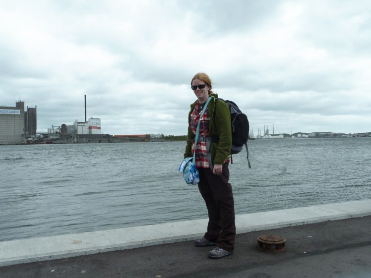 A timer selfie of me, full length, by the dockside in northern Denmark. The water is very high and very close. I'm wearing black hiking trousers, a red checked shirt under a green fleece and carrying my backpack and a small blue flowered handbag. Behind me on the other side of the water are some ugly industrial buildings.