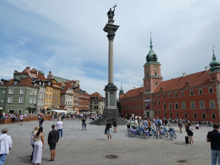 Castle Square, with a long three-storey orange-red building on the right, with a tower in the middle. In the middle of the square is a tall column with a Polish king on top. TO the right are a row of colourful merchant-style houses.