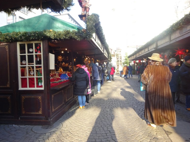 Inside the market. The streets are cobbled and lined on both sides by dark wooden stalls, with gold decorations at top and bottom and a long green wreath along the top dotted with lights. Dominating the scene is a woman in a straw hat and an ankle-length fur (or fur-like) coat.