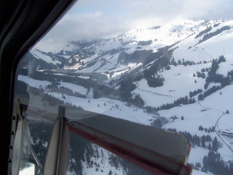 Flying up a snowy valley with a dark green river in the middle. The window of the helicopter is clearly visible.