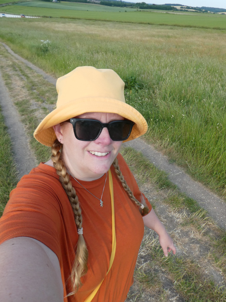 A selfie at Maiden Castle with a yellow bucket hat hiding my disgusting hair. I'm wearing an orange-brown t-shirt dress and sunglasses and holding the camera up high.