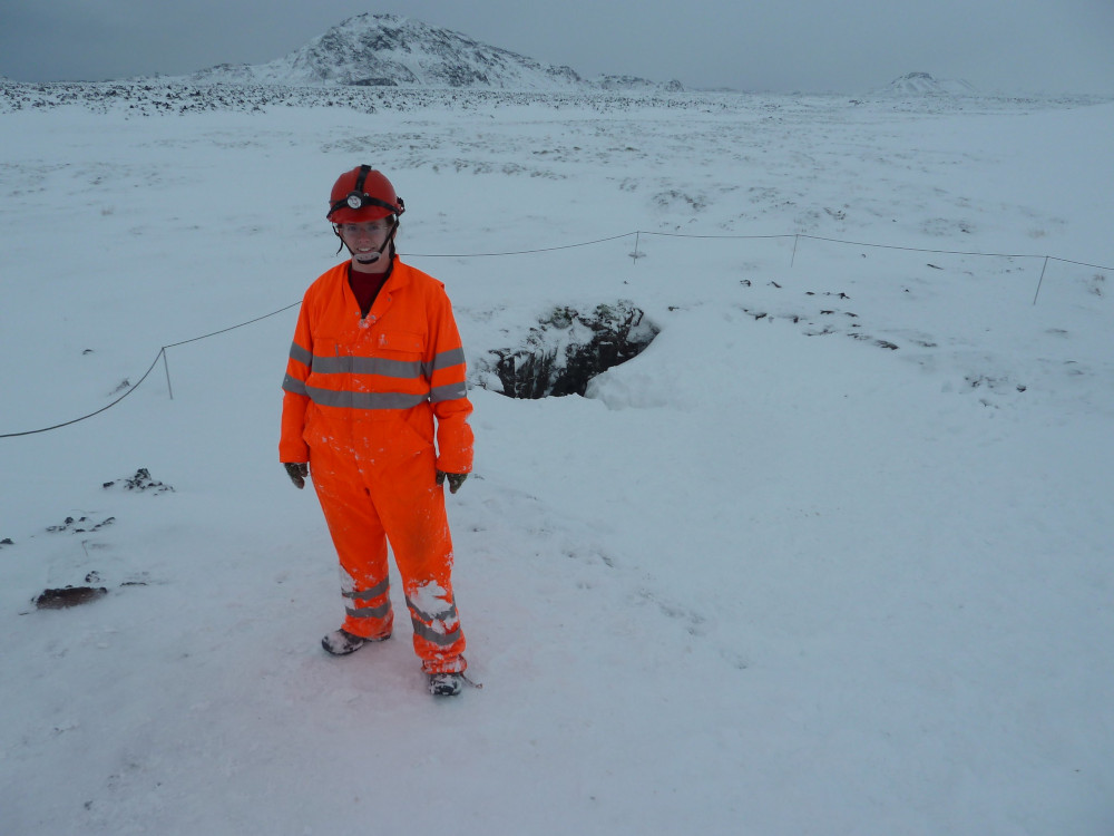 Me on a totally white background - well, there's a mountain but it's all thick snow and the sky is grey and you can't really see where the divide is. There's a hole in the ground behind me and I'm wearing a neon orange boilersuit with reflective stripes and a red helmet because I've just been caving.