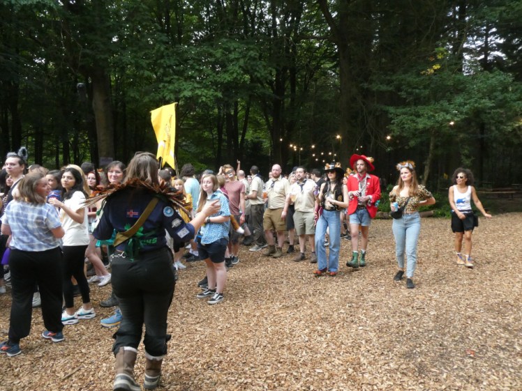 Friday evening parade - lots of people walking through the woods but the focus is on the Hawks Patrol Leader in the foreground with her back to the camera. She is wearing a navy blue shirt covered in badges, her old Brownie sash is sewn onto the shirt and she's wearing a collar of feathers to represent Hawk wings.