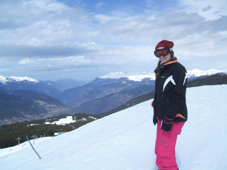 Me on the side of a snowy mountain. You can't see it because the picture cuts off around my knees but I'm on a snowboard, wearing bright pink ski trousers and a black jacket with orange and white accents, a red and black striped hat and goggles with reddish lenses.