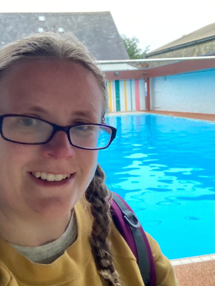 A selfie on the side of the pool. The water is a bright vivid turquoise blue behind me. My hair is plaited and wet from swimming, I'm wearing a yellow sweatshirt and carrying a blue and yellow kickboard