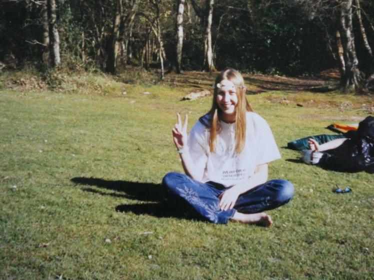 Catherine sitting on the grass wearing a daisy chain headband and making a peace sign.