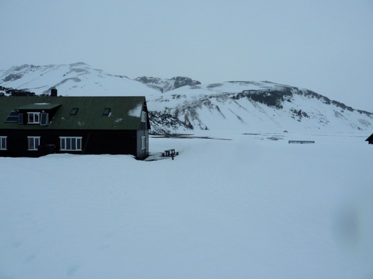 The campsite buildings, long wooden buildings with pointed roofs, in the snow. In the background is the mountain and the lava field and you can just make out the patch of warmth that is the hot spring.