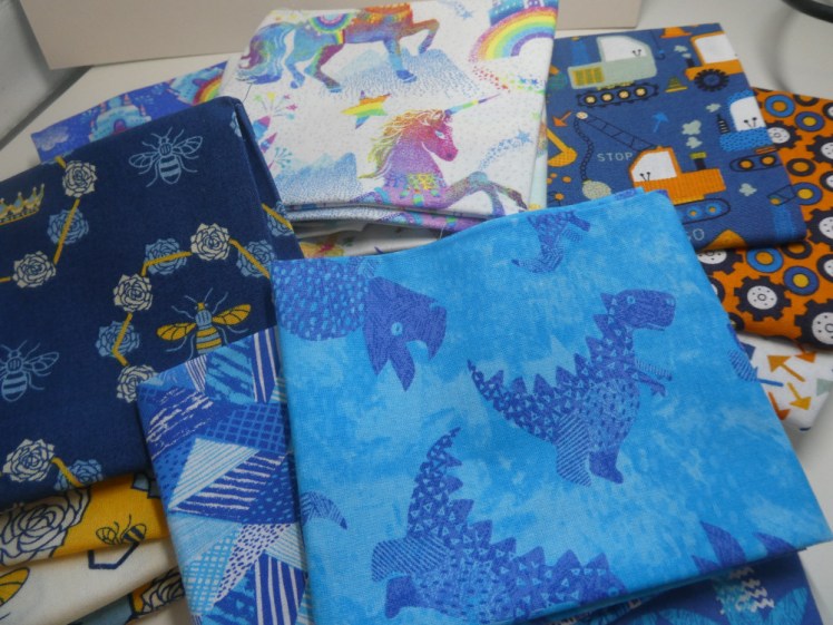 A pile of fat quarters folded into square. There's a pile of blue and yellow bee fabric, a pile of rainbows and unicorns, a pile of orange and blue diggers and cogs and on top, a pile of blue fabric with dinosaurs, palm trees and coordinating blue geometric shapes. I opted for the bee fabric.