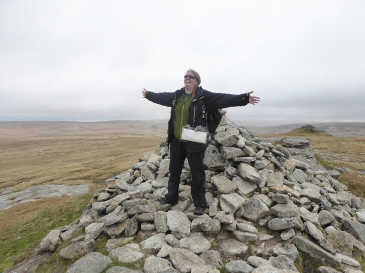 Me, in a green fleece and black rain jacket, on a pile of rocks marking the summit of High Willhays, the highest point in England south of the Lake District. My arms are outspread and I'm looking up at the grey cloudy sky in triumph.