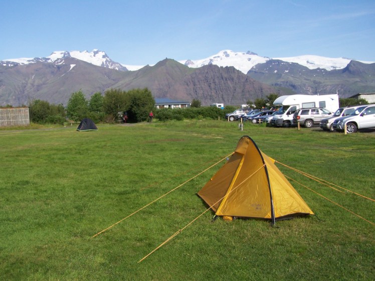 A small yellow tent, with a pole stretching from end to end rather than side to side. It has eight ludicrously long orange guylines stretched out. The grass is green and there's a perfect snow-capped mountain range behind it.